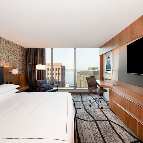 A modern hotel room with a large bed, TV, desk, chair, and a window with a city view. Walls are decorated, and furnishings are wooden.