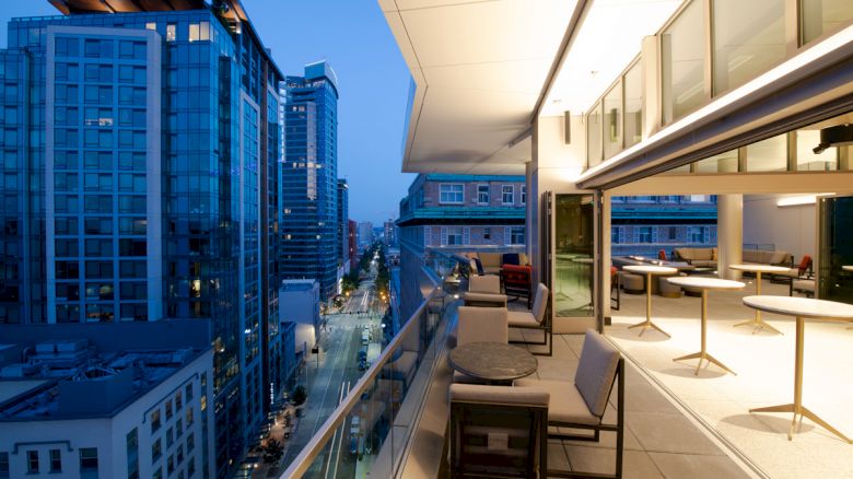 A modern balcony with seating overlooks a city street lined with tall buildings, illuminated by streetlights and city lights at dusk.