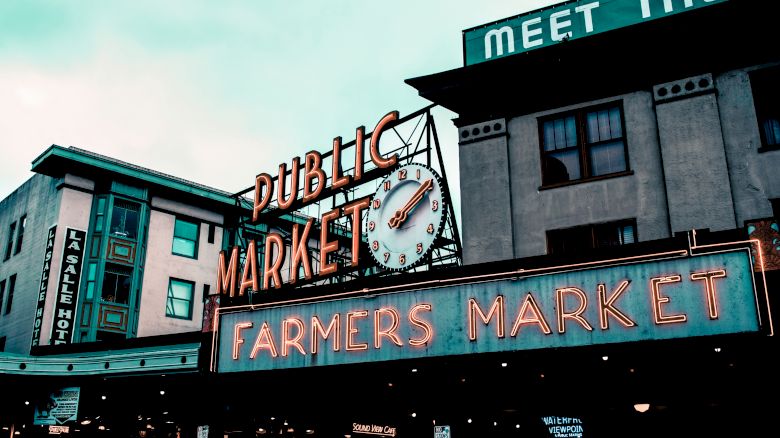 A large neon sign reads "Public Market" and "Farmers Market" with a clock above. Multiple buildings are in the background.