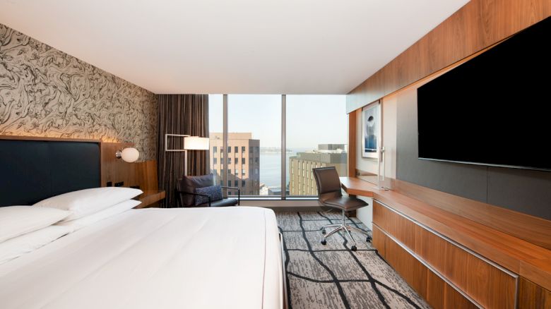 A modern hotel room with a large bed, desk, chair, TV, patterned walls, and a view of city buildings through large windows ends the sentence.