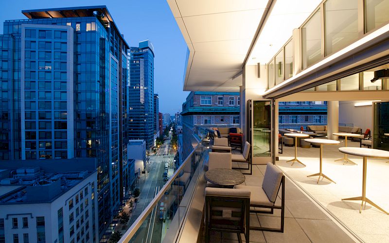 A modern cityscape at dusk with tall buildings, viewed from a stylish balcony featuring outdoor seating and tables under warm lighting.