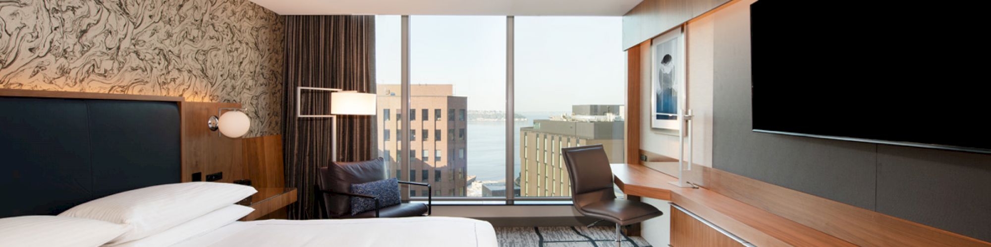 A modern hotel room with a large bed, TV, chair, desk, and a window offering a cityscape view. The decor is sleek and contemporary.