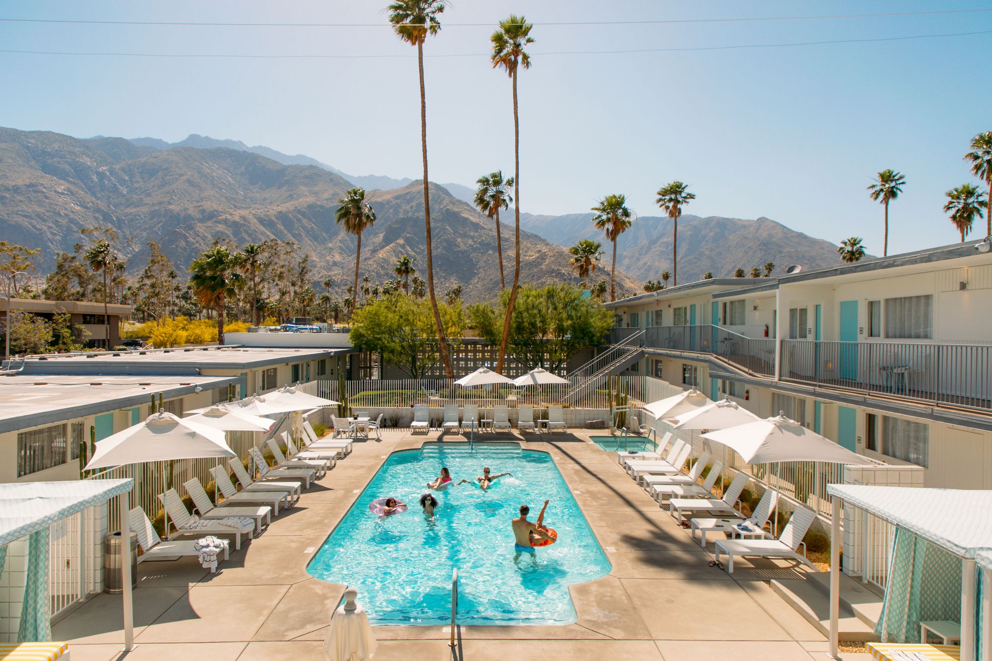 A sunny poolside scene at a retro-style motel with people swimming and lounging, surrounded by palm trees and mountains in the background.