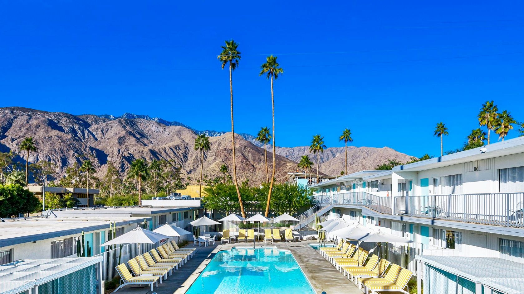 A sunny resort with a swimming pool, surrounded by yellow and white loungers, umbrellas, and palm trees under a clear blue sky and mountains.