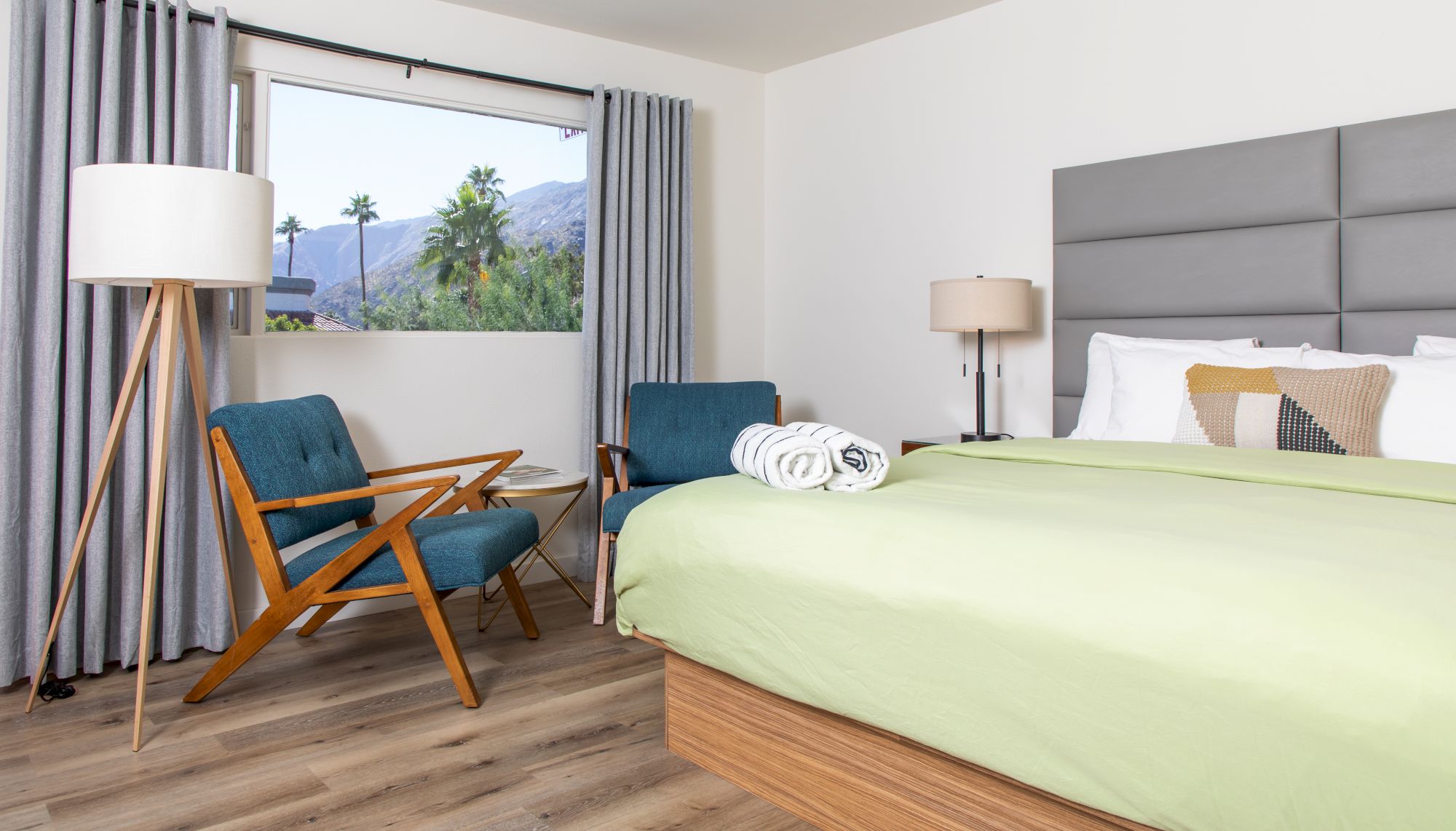 A modern bedroom features a green bed, chairs, a floor lamp, and a view of mountains through a window, with rolled towels on the bed.