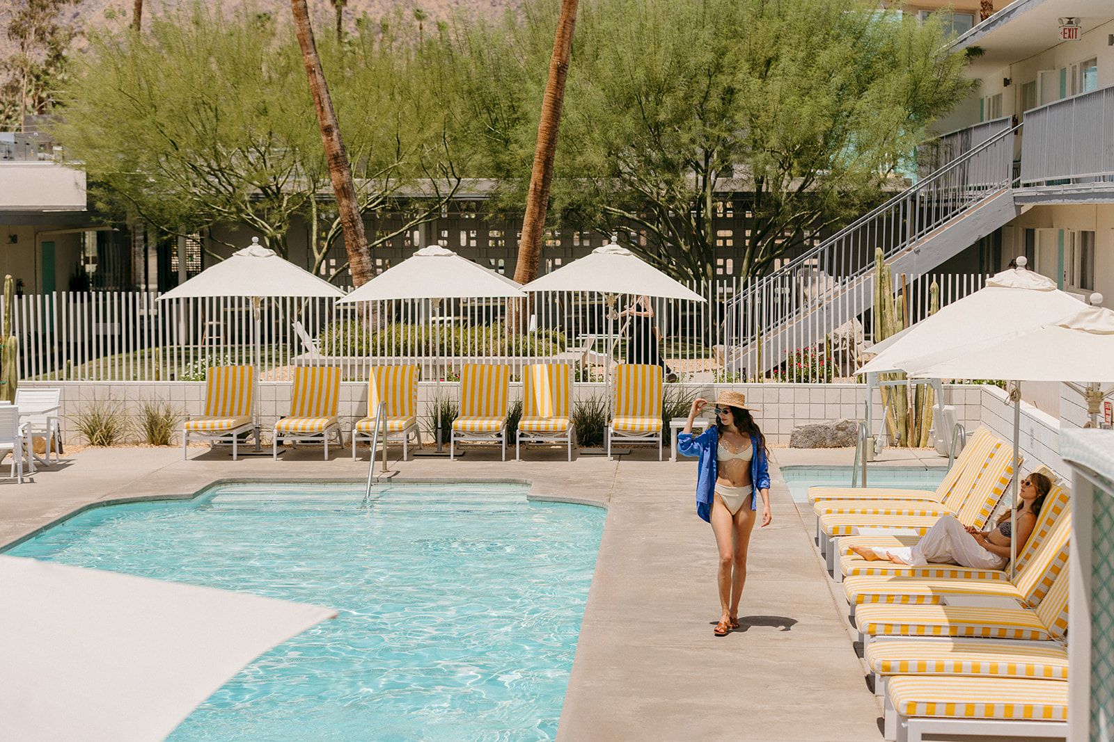 A poolside scene with people relaxing on lounge chairs, surrounded by umbrellas and greenery, with a modern building in the background.