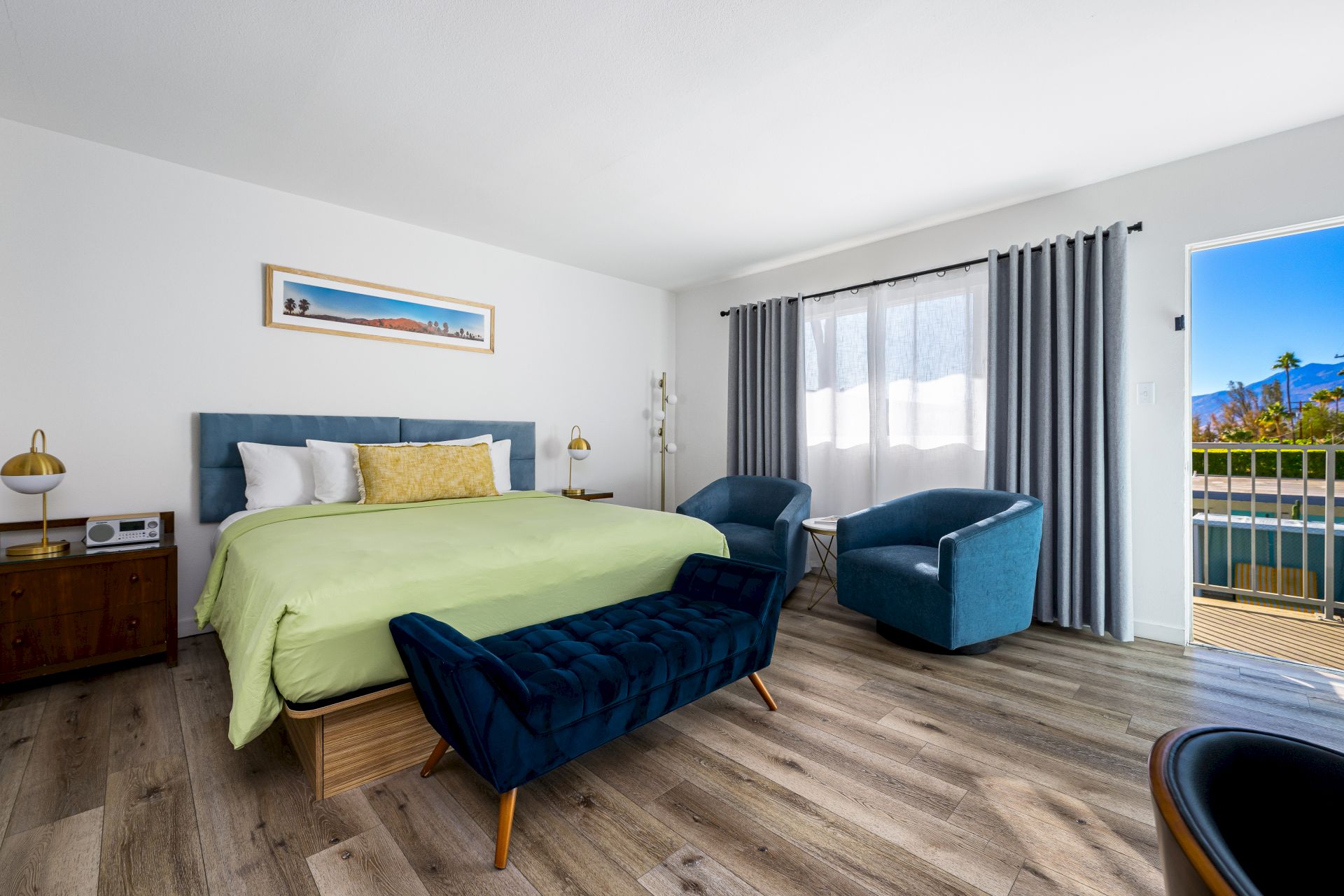 A modern bedroom with a green bedspread, blue seating, wood floors, and large windows with curtains. Art on the wall and a scenic view.