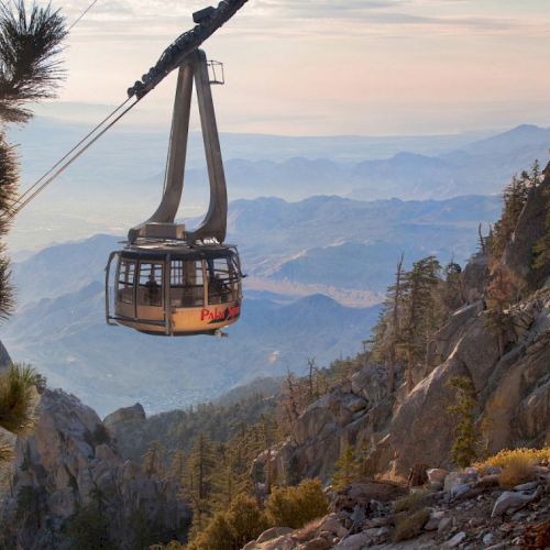 A cable car ascends over a mountainous terrain with scenic views of valleys and ridges under a cloudy sky.