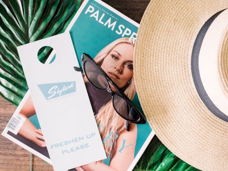 This image includes a Palm Springs magazine, sunglasses, a straw hat, and a "Freshen Up Please" door hanger on a tropical leaf backdrop, all on a wooden surface.