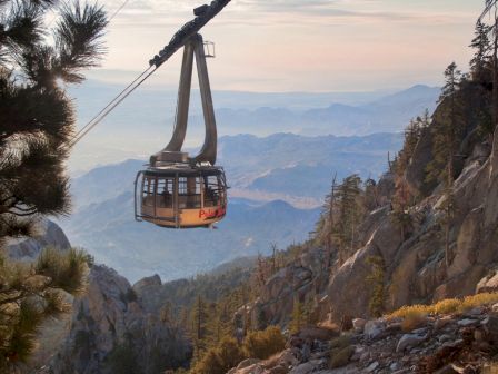 A scenic mountain view shows a tramway cable car suspended mid-air, surrounded by rocky terrain and pine trees, with a distant hazy horizon.